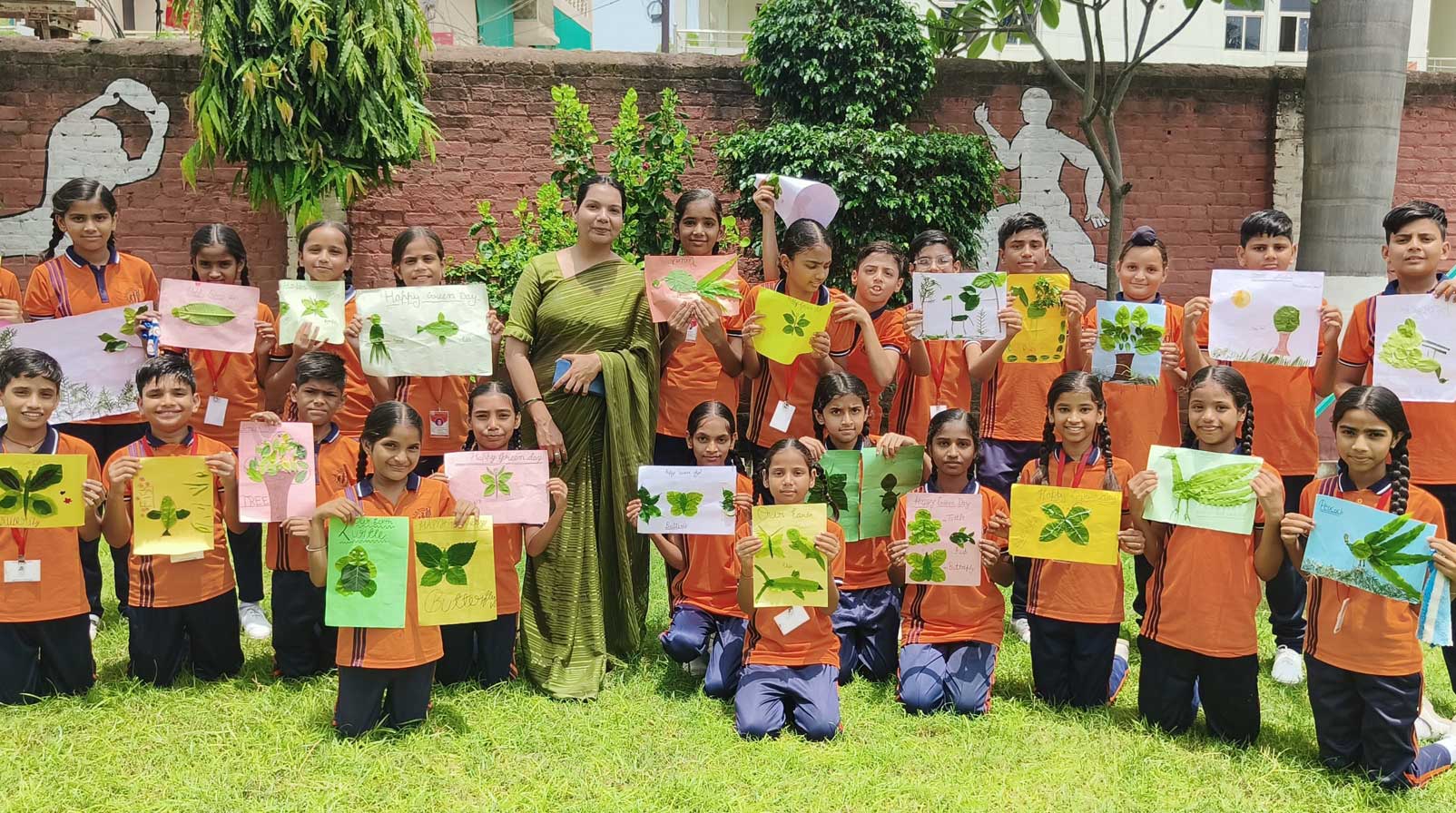 A teacher and many students in orange shirts proudly display their leaf-print art creations on green paper outdoors.