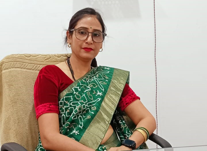 Woman, possibly the principal of SRDPS, wearing glasses and a green saree with a red blouse, seated in an office setting.