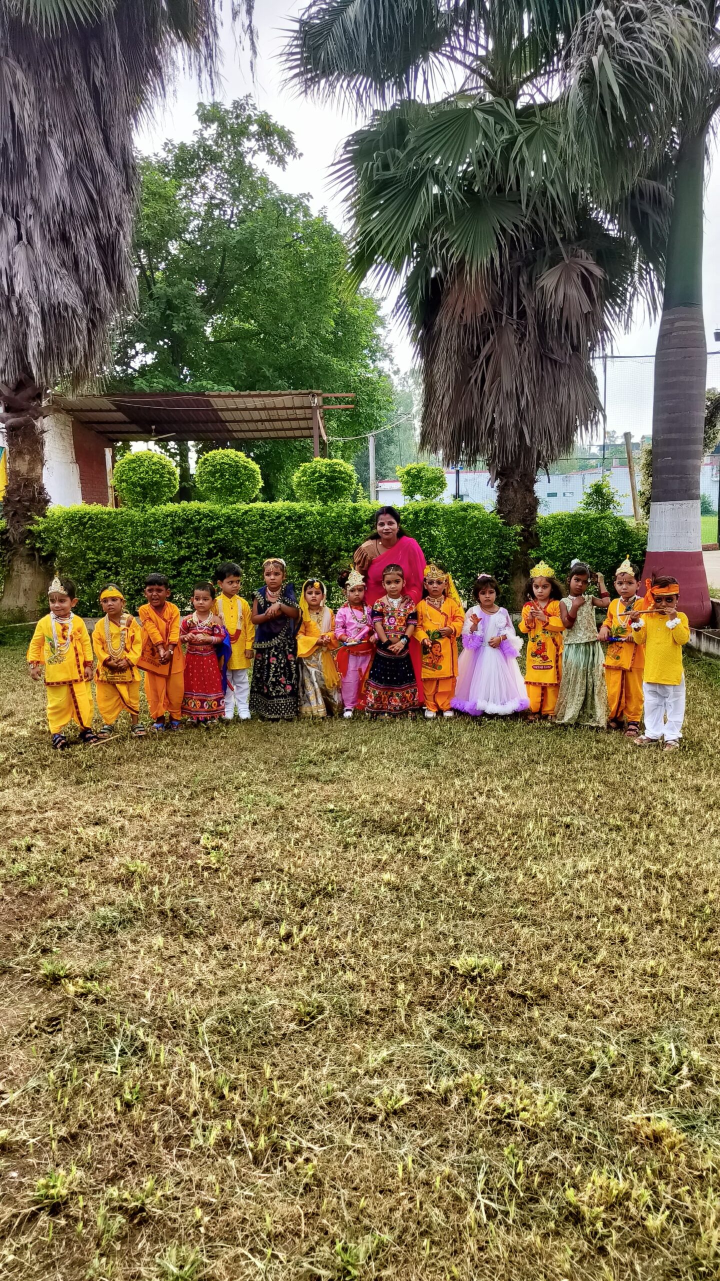 A female teacher poses with many young students dressed in colorful traditional Indian attire, standing on a grassy area outdoors.
