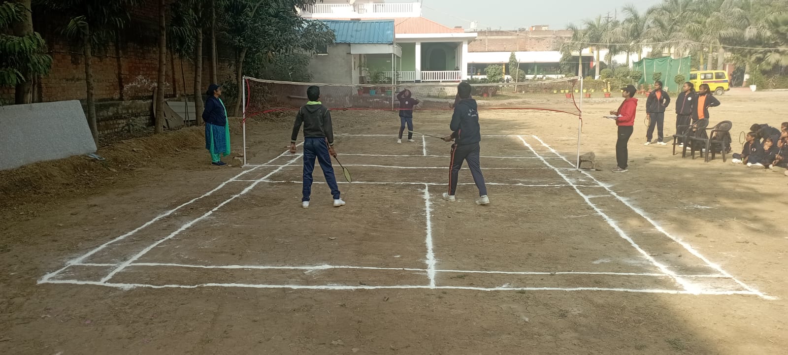 Students actively engaged in a badminton game on a dirt court marked with white lines, surrounded by trees and buildings.